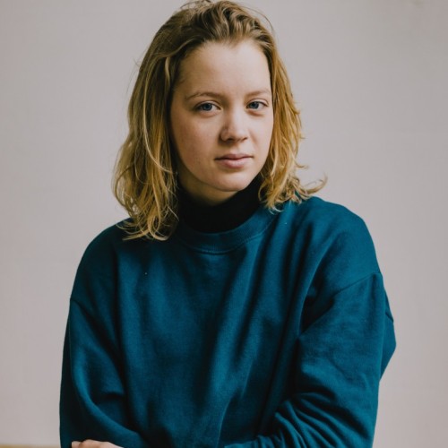 Carla steht vor einer grauen Wand. Sie hat einen dunkelgrünen Pulli an und lächelt halb in die Kamera. 