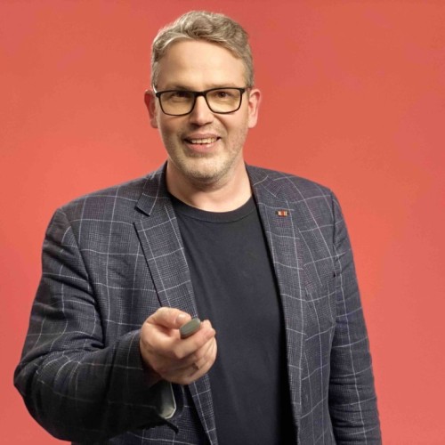 Mittelalter, leicht untersetzter Mann mit schwarzer Brille und Dreitagebart im blauen Sakko vor rotem Hintergrund, der auf einem Presenter klickt.