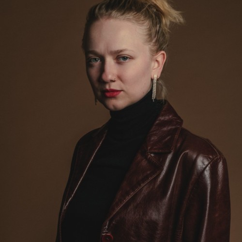 Mia Oberländer vor braunem Hintergrund. Sie trägt einen dunkelbraunen Ledermantel und roten Lippenstift.