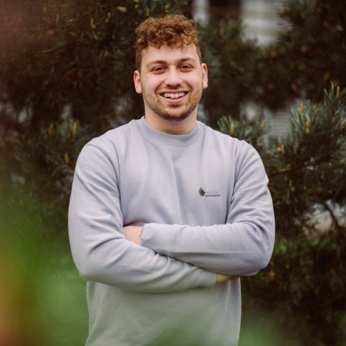 Auf dem Foto ist ein junger Mann zu sehen. Er hat rot-braune, kurze, lockige Haare, trägt einen fliederfarbenen Pullover vom Abenteuer Regenwald e.V. und lächelt fröhlich in die Kamera. Seine Arme sind vor dem Überkörper verschränkt. Im Hintergrund befindet sich ein Baum.