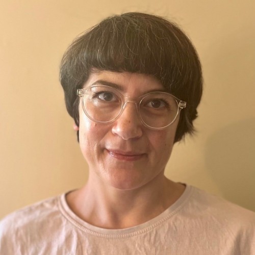 Frau mit dunkelbraunem kurzen Bob und heller Brille, hellem T-Shirt vor hellem Hintergrund