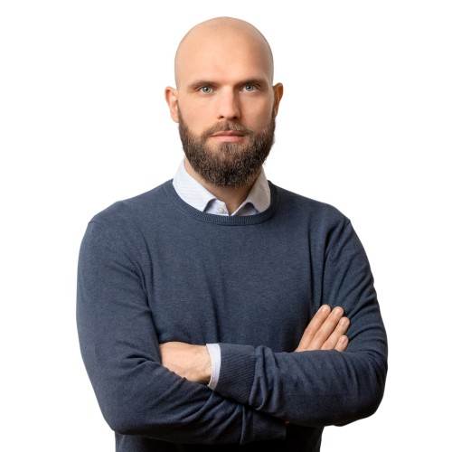 Porträt von Hannes Schwarm, ein Mann mit Glatze und Vollbart, vor weißem Hintergrund. Er trägt ein hellblaues Hemd unter einem dunkelblauen Pullover. Die Arme sind vor der Brust verschränkt. Der Gesichtsausdruck ist neutral bis ernst, der Blick ist direkt in die Kamera gerichtet.