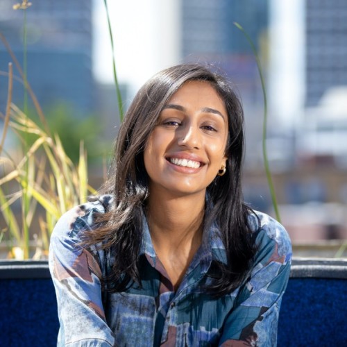 Image of a brown woman with dark hair, wearing a blue shirt. Background is of a cityscape 