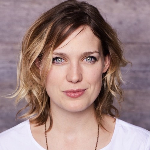 Cori ist Mitte 30 und Irin. Sie hat hellblaue Augen und dunkelblondes Haar. sSie trägt ein weißes T-shirt.