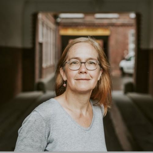 Das Bild zeigt die Journalistin Heike Kleffner in einem grauen Pulli vor einer Toreinfahrt.