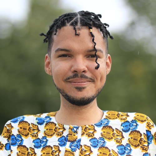Portrait of smiling Djamal wearing a shirt with a colourful floral motif, and a short beard. One of the fresh twists hangs in his face. 