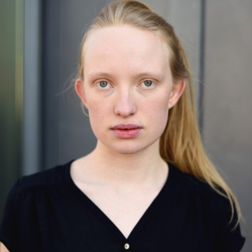 Das Portrait einer weißen, blonden jungen Frau mit Zopf vor einem grauen Hintergund. Sie trägt eine schwarze, kurzärmlige Bluse und ihr halber Oberkörper ist zu sehen.