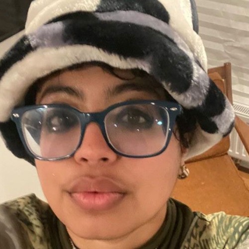 A brown skinned person witu a grey, black and white faux fur hat with glasses and kohl-adorned eyes.