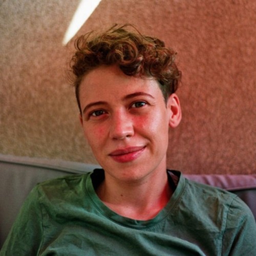 Das Bild zeigt ein frontales Portraitfoto von Cin. Cin guckt entspannt nach vorne, der Kopf ist leicht nach links geneigt. Cin hat kurze, braune Locken, volle Lippen, leichte Lachfältchen um die Augen und ist weiß. Cin trägt ein Tshirt in einem mittleren Grünton. Im Hintergrund ist eine beige Hauswand und ein graues Polster an die Cin angelehnt ist. Ein Sonnenstrahl bricht durch den oberen Bildrand links an Cins Kopf vorbei, das Bild hat einen leicht orange-roten Einschlag und wirkt sommerlich.