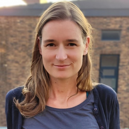 Foto von Kristina Richter im freien vor einer Backsteinfassade im Hintergrund. Offenes Haar, dunkelblaue Kleidung, Lächeln.