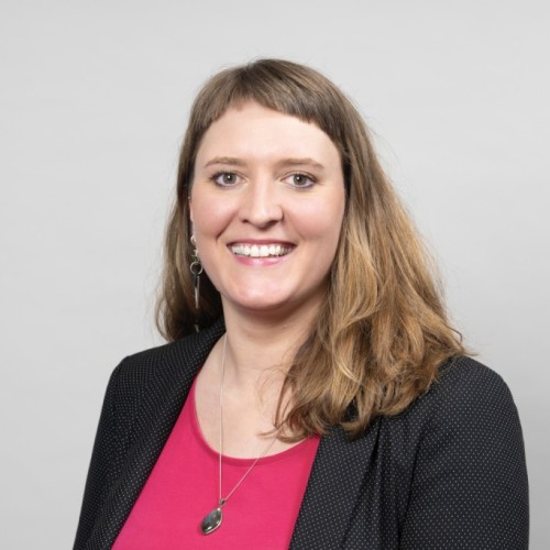 Das Foto zeigt das Portrait einer Frau, die vor einem weißen Hintergrund steht und in die Kamera lächelt. Sie trägt einen schwarzen Blazer und eine pinke Bluse.