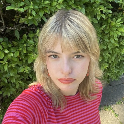 Auf dem Bild sieht man eine junge Frau mit Blonden Haaren und einen Rot-Pink gestreiftem T-Shirt vor einem grünen Hintergrund. 