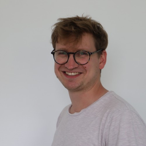 David Hartmann mit einer runden Brille und einem hellen T-Shirt lächelt in die Kamera. Der Hintergrund ist schlicht weiß, und das Bild zeigt eine freundliche, entspannte Ausstrahlung.