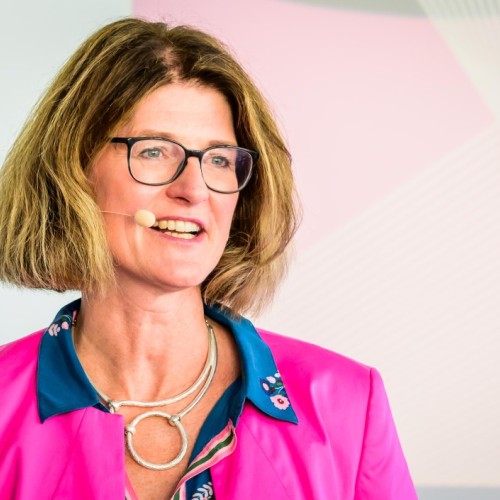 Karen Funk ist sichtbar vom Kopf bis zum mittleren Oberkörper, sie trägt ein Headset und ein pinkfarbenes Sakko.