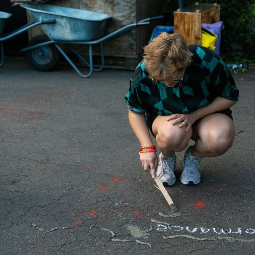Iz, a white nonbinary person with short blonde hair who is wearing a green shirt and sunglasses, is kneeling on the ground during a performance of their arts-design duo MELT. They are using a stick to pour concrete into a crack on the floor.