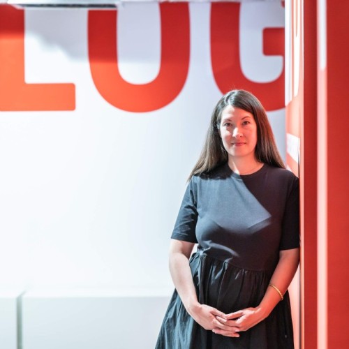 Maureen Ekizoglu lehnt mit ihrer linken Schulter gegen eine rote Wand. Sie lächelt freundlich in die Kamera.