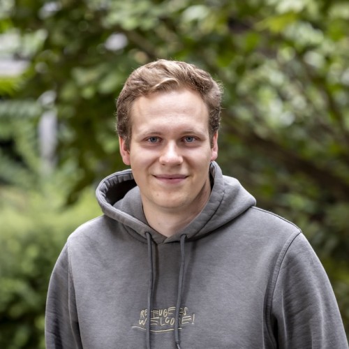 Ein junger Mann mit kurzem, hellbraunem Haar steht im Freien vor einem unscharfen, grünen Hintergrund aus Büschen und Bäumen. Er trägt einen olivgrünen Kapuzenpullover und blickt lächelnd in die Kamera. 