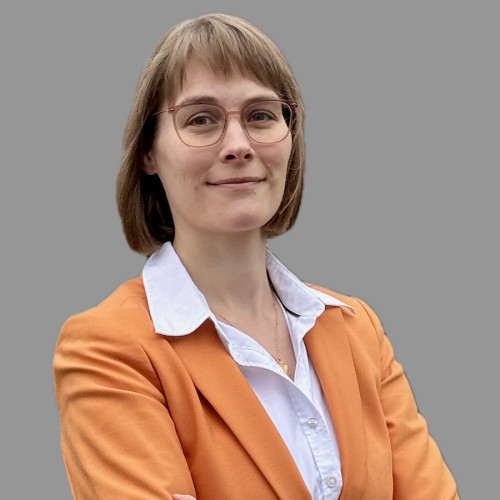 Blonde Frau mit Haaren bis kurz unter die Ohren und Pony trägt eine Brille. Schaut in die Kamera. Sie trägt eine weiße Bluse und Orangen Blazer. Ihre Schultern und ein kurzer Teil des Oberkörpers ist zu sehen.