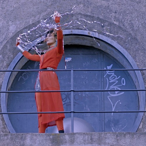 A portrait of Jessika Khazrik standing next to a silver sphere against a concrete backdrop with a circular gate, holding with her raised arms a web of scintillating threads