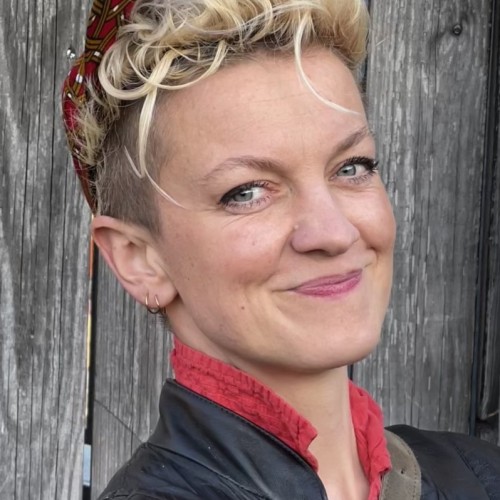Lillian Rosa, kurze blonde Haare und rote Käppi, steht vor einer Holzwand und lächelt in die Kamera