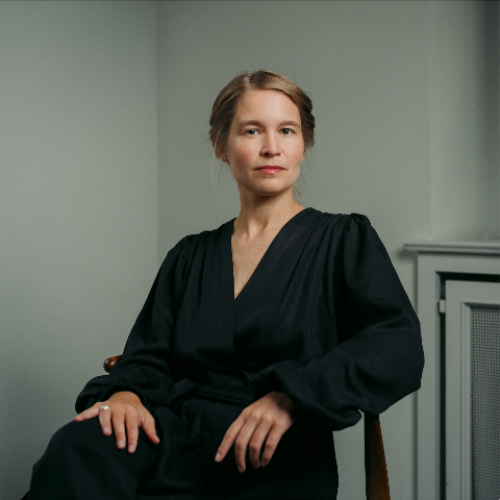 Eine Frau mit blondem Haar, das zu einem schlichten Knoten zurückgesteckt ist, sitzt auf einem Stuhl und blickt ruhig und selbstbewusst in die Kamera. Sie trägt ein elegantes, schwarzes Kleid mit tiefem V-Ausschnitt und langen, leicht gepufften Ärmeln. Ihre Hände ruhen entspannt auf ihren Knien, einer ihrer Finger ist mit einem Ring geschmückt. Der Hintergrund ist minimalistisch und in neutralen Farbtönen gehalten.