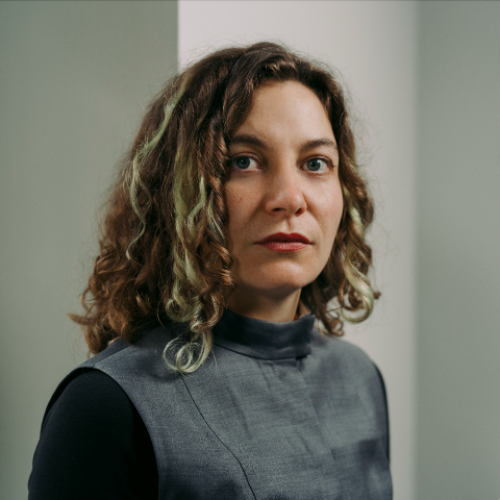 Eine Frau mit lockigem, schulterlangem Haar und grünen Strähnen blickt ernst in die Kamera. Sie trägt ein hochgeschlossenes, grau-blaues Oberteil über einem schwarzen Langarmshirt. Ihr Ausdruck ist ruhig und selbstbewusst. Der Hintergrund ist minimalistisch mit neutralen Farbtönen.