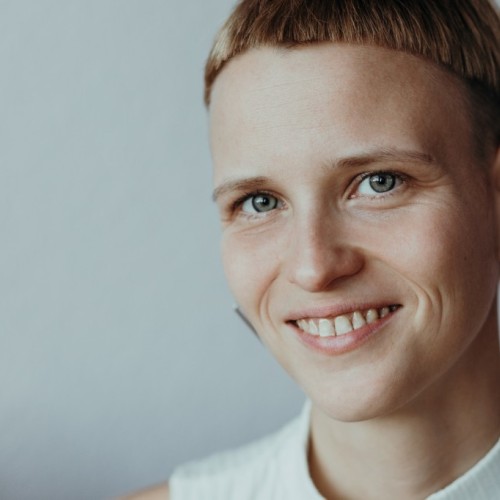 Manuela hat kurze blonde Haare, dreieckige Ohrringe und lächelt in die Kamera.