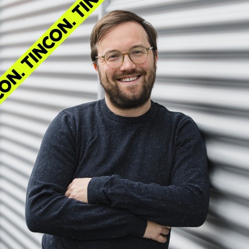 Junger, lächelnder Mann trägt Brille, Dreitagebart und einen Strickpullover