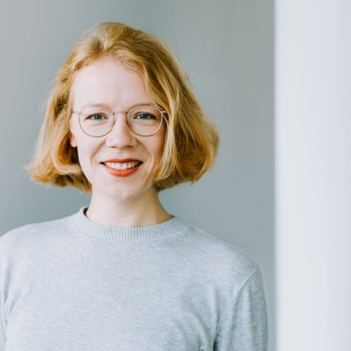 Auf dem Foto abgebildet ist eine Frau mit kinnlangen blonden Haaren, die in die Kamera blickt. Sie steht vor einem hellen Hintergrund und trägt ein hellgraues Oberteil.
