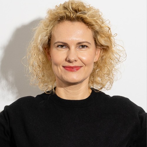 Eine Frau mit hellen blonden Locken und rotem Lippenstift schaut gerade und erwartungsfroh in die Kamera.