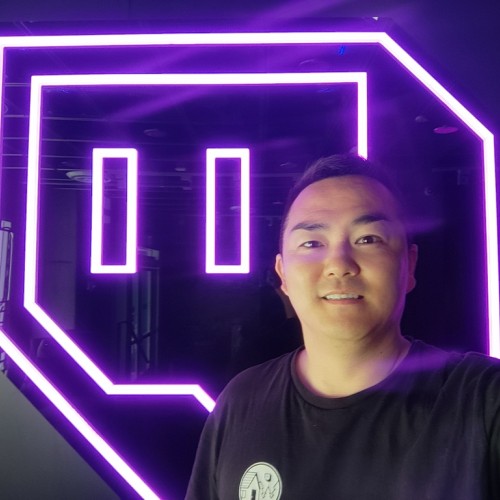 A picture of Mitsuo, smiling, short hair and a black shirt. In the background the Twitch logo in neon style.