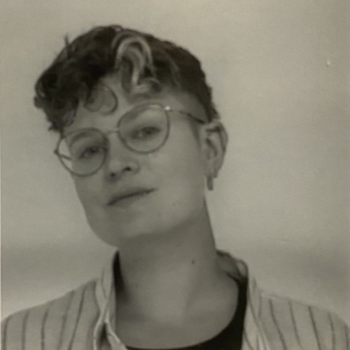 Eine Person mit Brille, gestreiftem Hemd und kurzen Locken schaut mit leicht schiefgelegtem Gesicht in die Kamera.