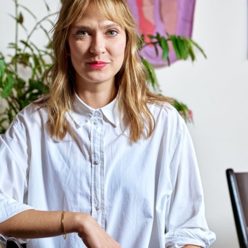 Person with long blond hair sitting on a chair and looking straight into the camera. 