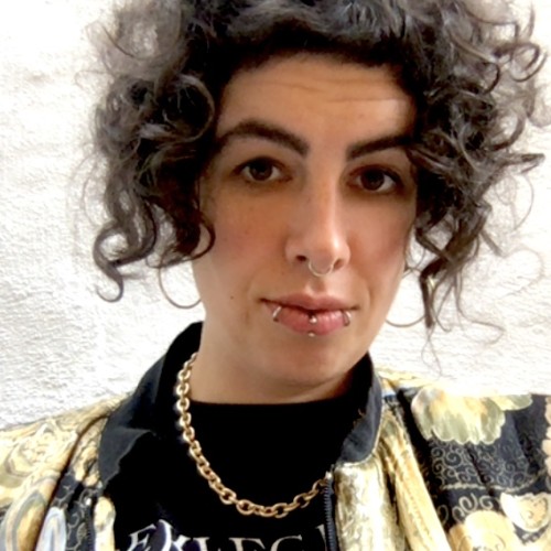 Nushin has rather light skin and brown chin length curls, weards a black and gold jacket and a necklace