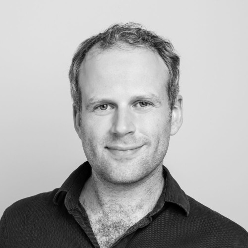Black and white headshot of Oliver wearing a dark shirt against a plain background. Oliver is a white mid 30s man with dark hair