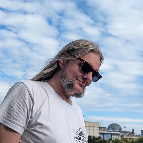 Ein Portrait von Olli im High Ground Shirt auf der Hanfparade 2023, mit Schäfchenwolken vor blauem Himmel im Hintergrund.