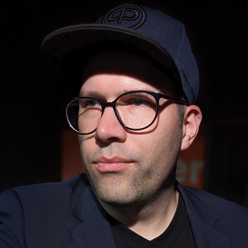 Ein Portrait-Foto von Thomas Riedel mit starken Kontrasten. Thomas blickt nach Link. Er trägt eine dunkelblaue Schildmütze mit einem aufgestickten typographischen Motiv, das wir ein verschmolzenes kleines q mit einem großen P ähnelt. Er trägt dazu ein fast gleichfarbenes dunkelblaues Sacko und ein schwarzes T-Shirt mit V-Ausschnitt. Seine schwarze Brille hat eine fast runde Form und wirft Schatten auf sein Gesicht. Zwei-Tage-Bart.