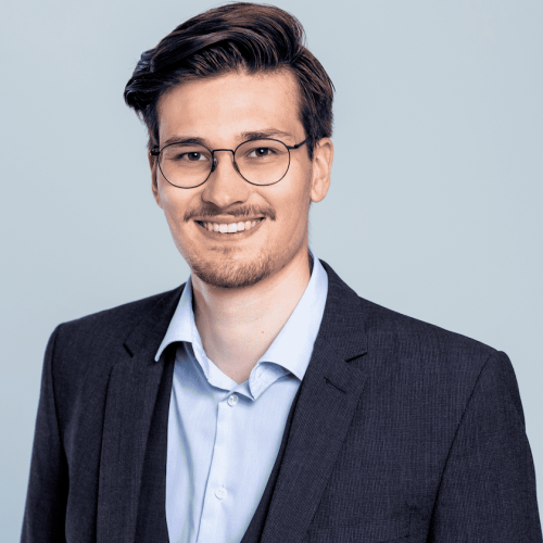 Junger Mann im Dreiteiler, mit Bart, Brille und braunen Haaren.
