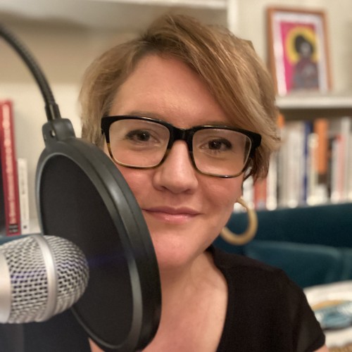 A portrait of a white woman, mid-30s, with short dark blonde hair. She is wearing a black tshirt and glasses with dark thick rectangular frames. She is smiling and sitting behind a microphone with a pop guard and in front of a bookcase.
