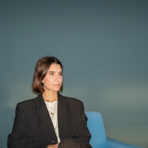 White woman with brown hair wearing a black blazer over a white shirt sitting on a blue armchair.