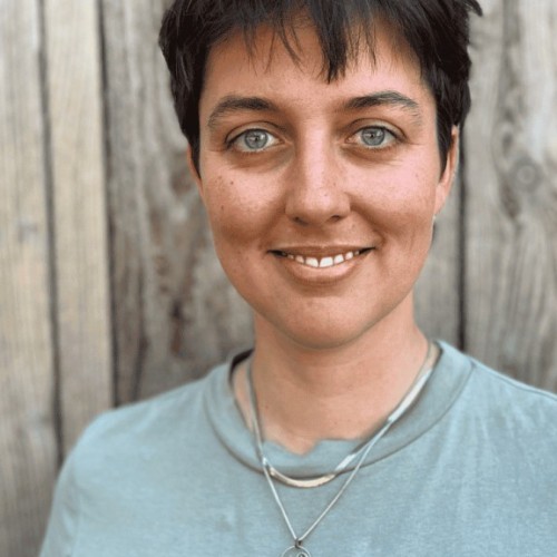 Profilbild Johanna Balsam, lächelnde Frau im grünen T-Shirt, mit kurzen braunen Haaren vor einer Holzwand stehend