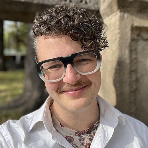 Ren smizes at the camera, they have short brown curly hair, big glasses and a queer looking mustache. Its sunny and they're outside.