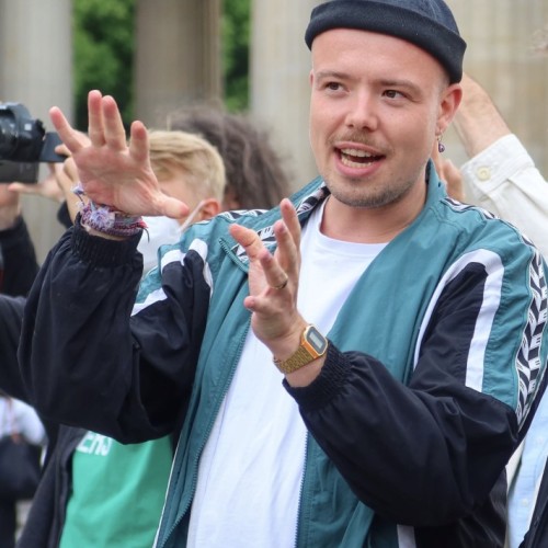 Robin Elias Alef steht draußen vor einem Gebäude mit großen Säulen. Er trägt eine blaue Mütze, eine türkis-schwarze Sportjacke mit weißen Streifen, ein weißes T-Shirt und eine Digitaluhr. Er spricht gestikulierend mit erhobenen Händen, als würde er etwas erklären oder betonen. Im Hintergrund sind andere Menschen zu sehen, einige mit Kameras oder Masken. Die Atmosphäre wirkt lebendig und engagiert.