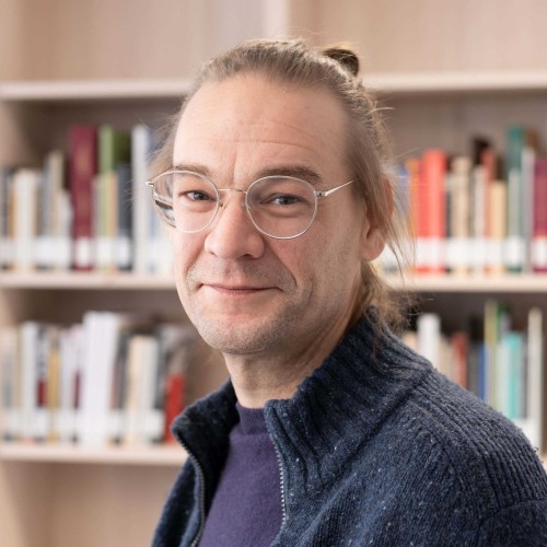 Lächelnder Typ vor Bücherwand mit Brille und man-bun.