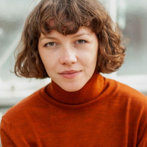 Auf dem Bild ist Saskia zu sehen, die direkt in die Kamera schaut. Sie trägt einen orangenen Pullover, ihre Haare sind offen, gelockt und etwa kinnlang und sie hat einen Pony.