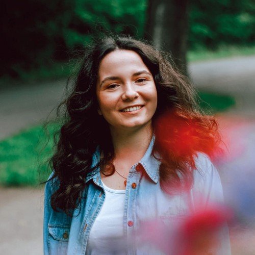 Zusehen ist eine Frau mitte zwanzig mit dunkelbraunen langen gewellten Haaren. Sie lacht in die Kamera. Hinter ihr ist in der Unschärfe eine Baum zu erkenne. Sie trägt ein weißes Top mit blauem Jeanshemd darüber. Sie ist leicht geschminkt.