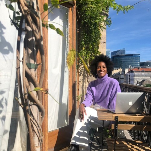 Black woman with an afro, smiling, wearing a purple pullover and white pants