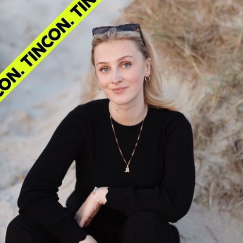 Frau mit blonden Haaren, schwarzer Sonnenbrille auf dem Kopf und schwarzem Pullover sitzt im Sand