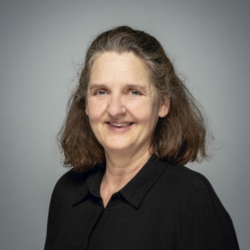 Porträtfoto einer Frau mit langem braunem Haar. Sie trägt eine schwarze Bluse und lächelt leicht in die Kamera. Der Hintergrund ist grau.