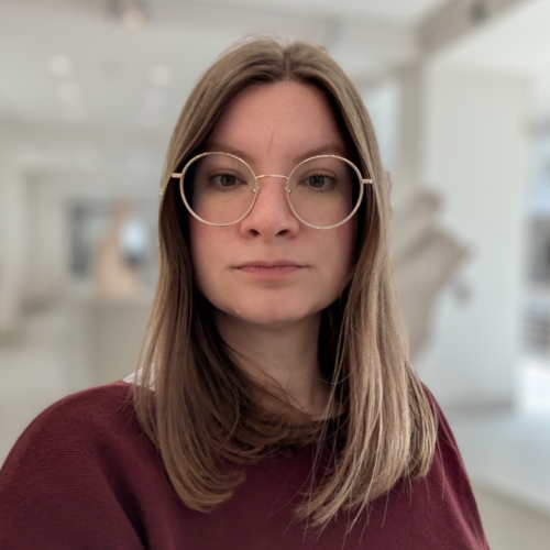 Portrait einer weißen Frau. Sie hat blonde, glatte Haare und trägt eine runde, goldene Brille.